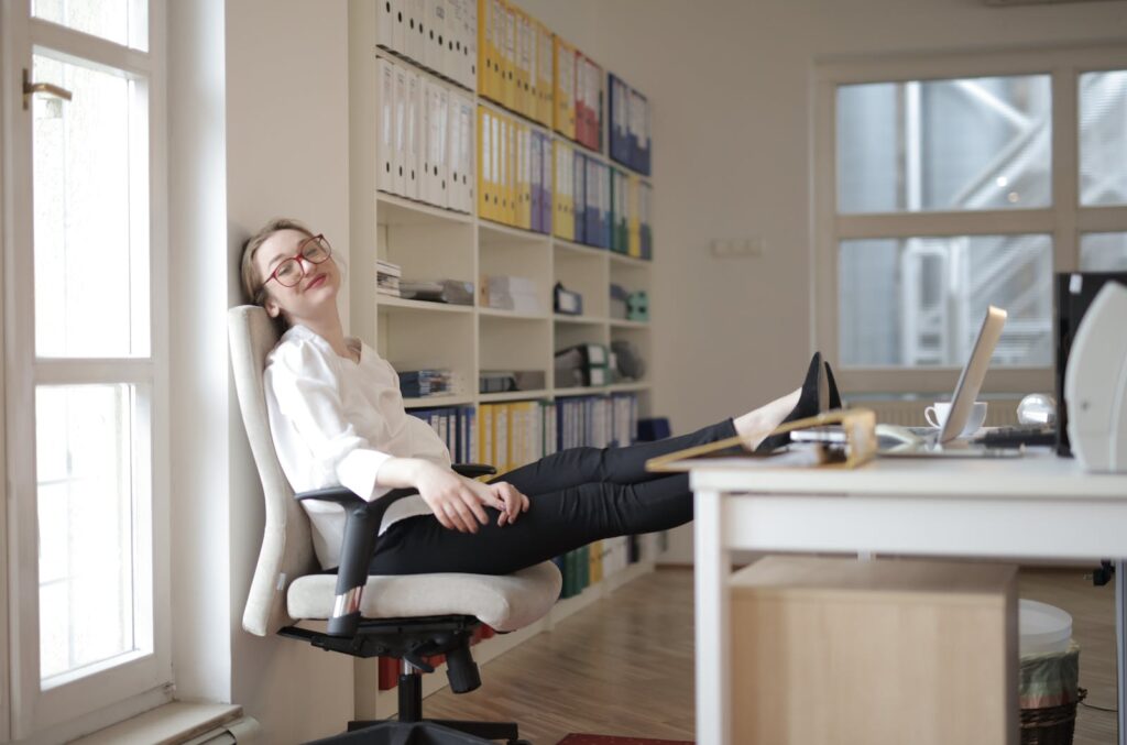 Side view of content female employee wearing formal clothes and eyeglasses sitting on chair with crossed feet on table and chilling during work with closed eyes in modern office